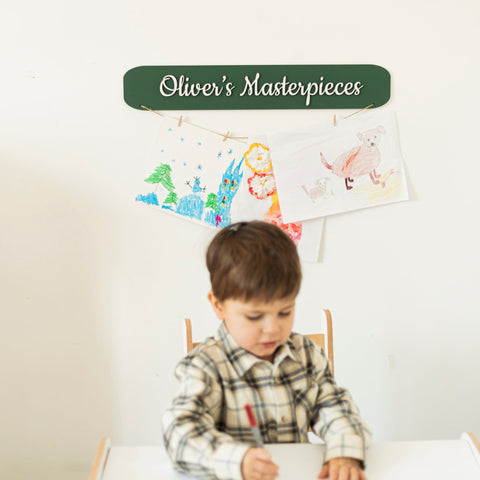 Child sitting at a table with drawings on the wall above, labeled 'Oliver's Masterpieces'.