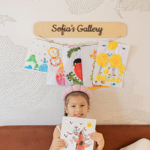 Child holding a drawing in front of a wall with children's artwork and a sign that reads 'Sofia's Gallery'.