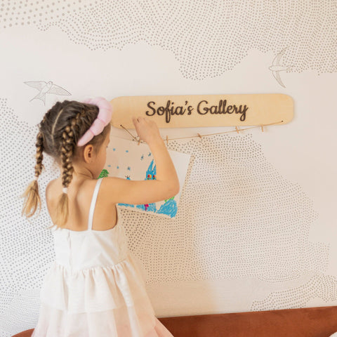 Child with braided hair and pink headband holding a wooden board with 'Sofia's Gallery' on it against a textured wall.