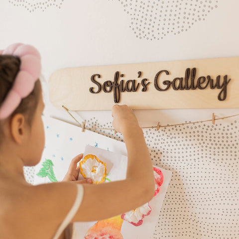 Child interacting with a wooden art display named 'Sofia's Gallery' on a textured wall.