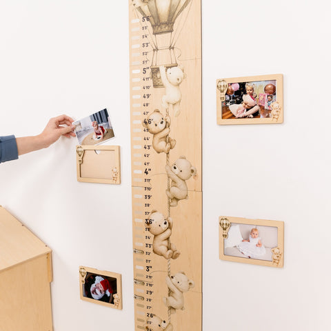 Wooden growth chart with teddy bear markers and photo frames on a white wall.