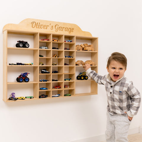 Child standing next to a wooden toy car shelf with 'Oliver's Garage' on a white wall.