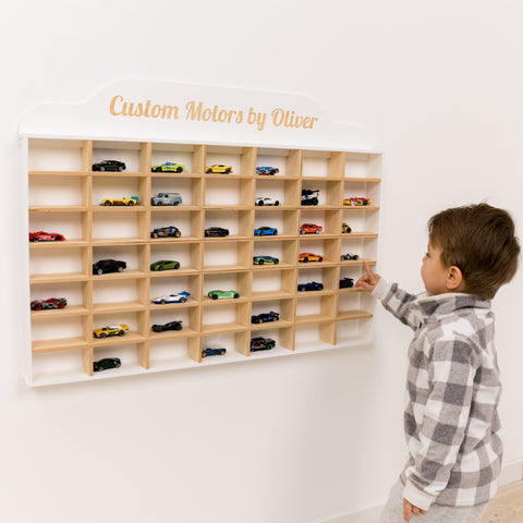 Child interacting with a wooden wall display of toy cars labeled 'Custom Motors by Oliver'.